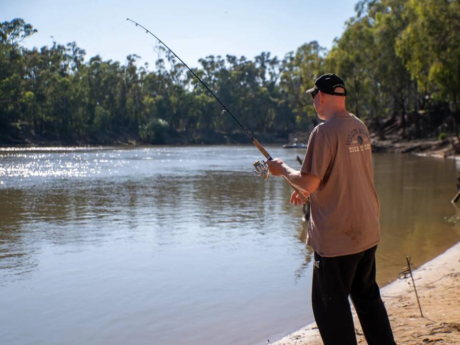 A person fishing by a calm river with trees in the background.