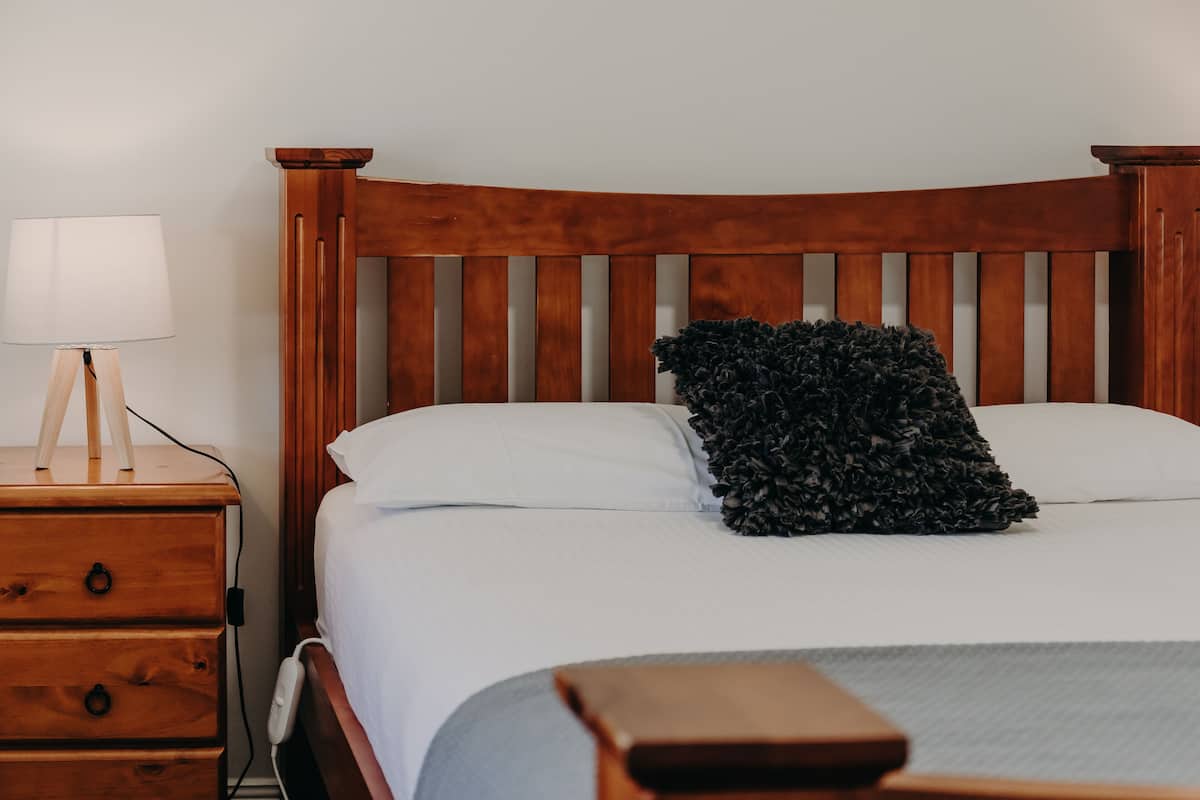 Ein ordentlich bezogenes Bett mit Holz-Kopfstück, ein Nachttisch mit Lampe und ein dunkel strukturiertes Kissen.