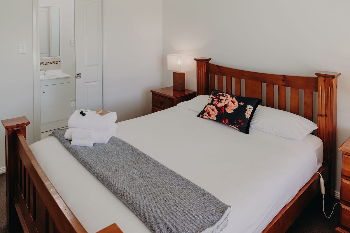 Ein ordentlich bezogenes Bett mit einer grauen Tagesdecke, einem hölzernen Kopfteil und einem floralen Kissen. Ein Nachttisch mit Lampe und einer kleinen Schublade ist ebenfalls vorhanden. Eine weiße Tür führt zu einem Badezimmer.