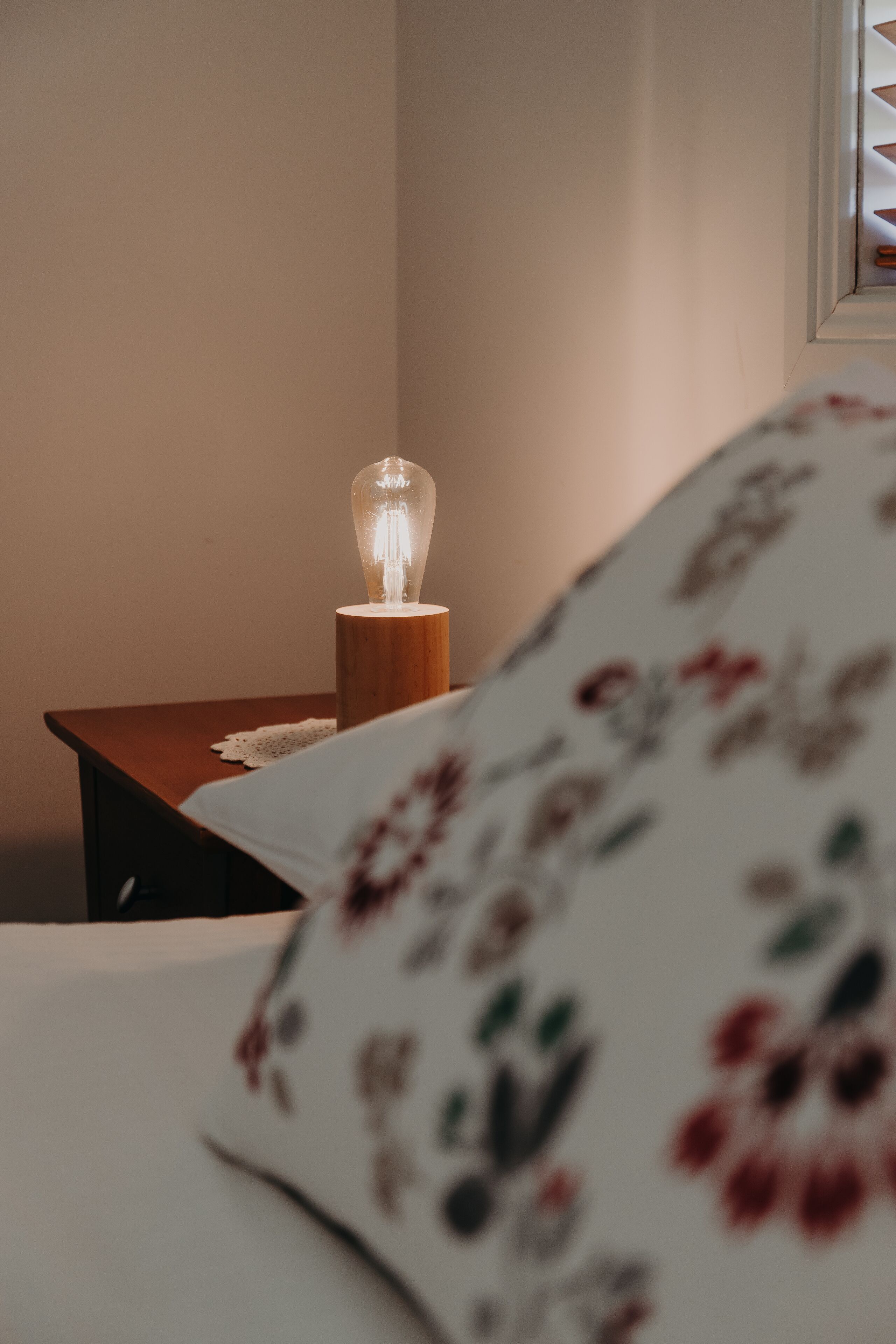 A bed with a floral-patterned blanket, a bedside table with a lamp, and a framed picture on the wall.
