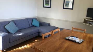 A living room with a sofa, two framed pictures on the wall, a television on a shelf, and a dining table with chairs.