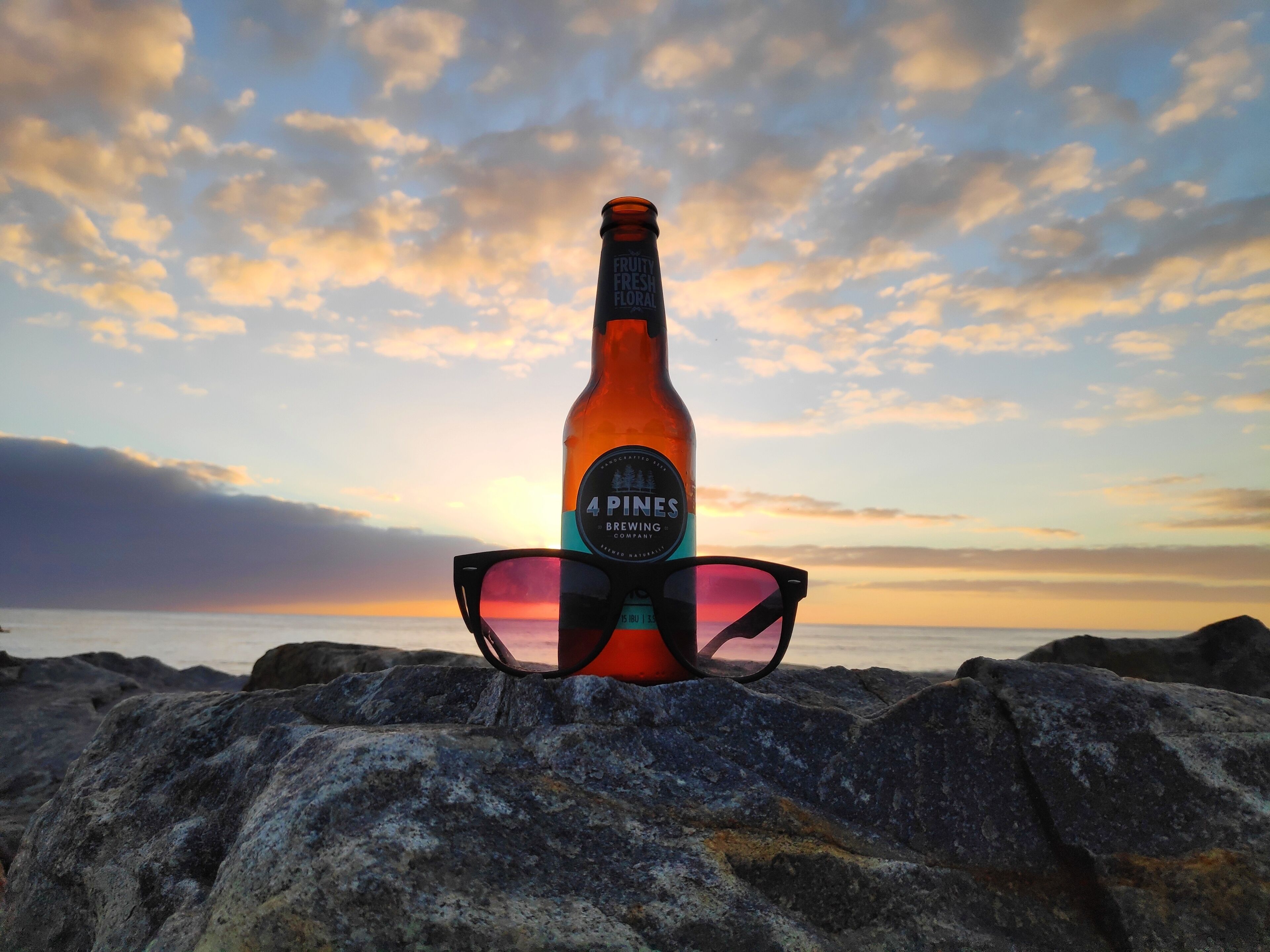 A bottle of 4 Pines Brewing IPA and a pair of sunglasses on a rocky surface with a sunset in the background.