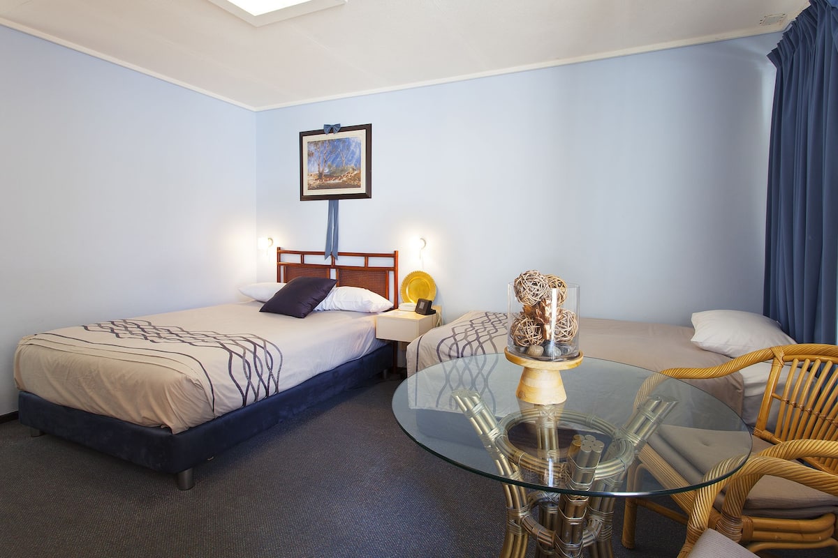 Una habitación de hotel con dos camas, una mesa de vidrio, sillas de mimbre y un cuadro en la pared.