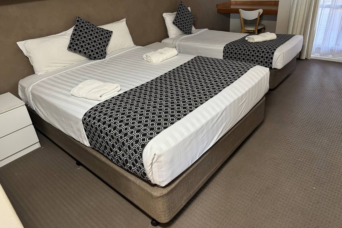 A hotel room with two beds, each with a patterned bedspread and white pillows. There is a small desk with a chair and a window with curtains.