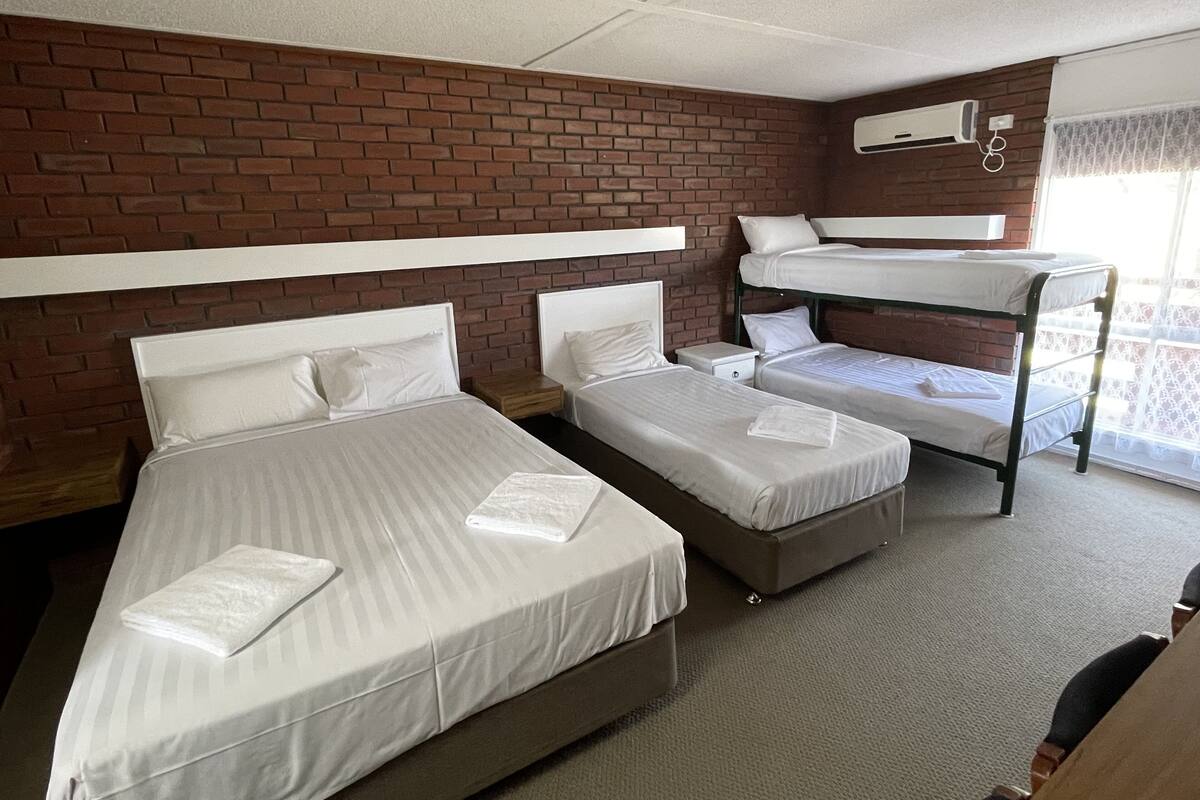 A hotel room with three beds, a brick wall, a window with a curtain, and a ceiling fan.