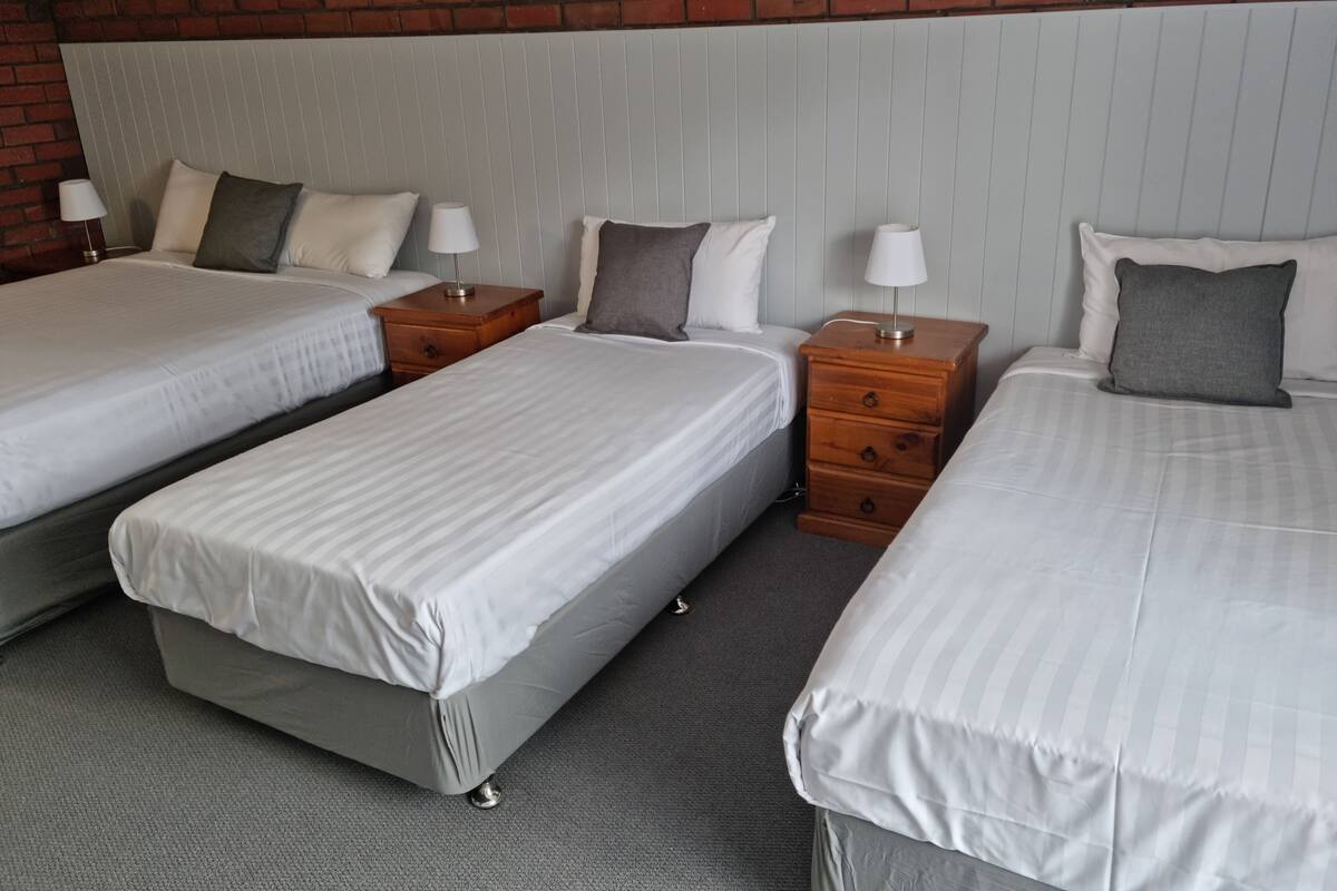A hotel room with three beds, each with white bedding and grey pillows. There are wooden nightstands with lamps and drawers. The room has a brick wall and a white headboard.