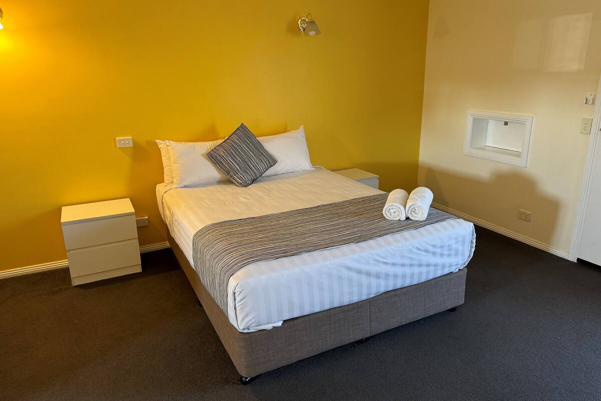 A neatly made bed with white and striped bedding, a grey upholstered headboard, and two rolled towels on the foot of the bed.