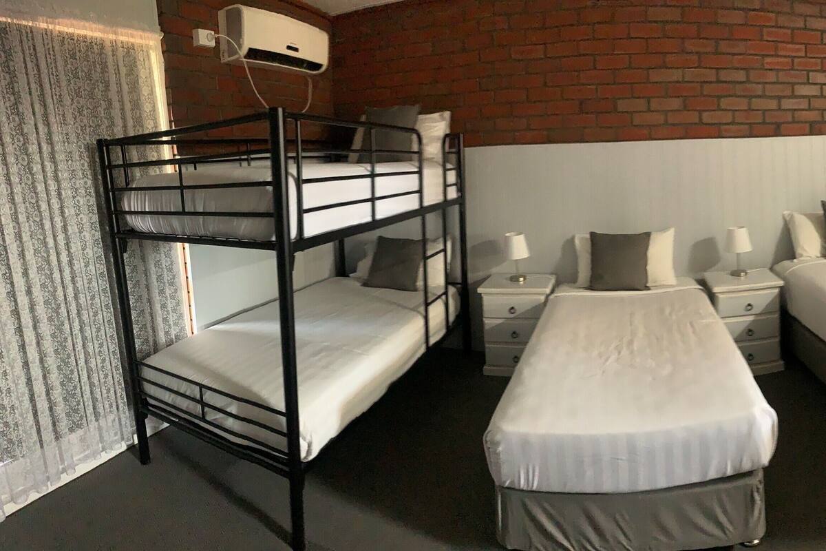 A room with a bunk bed, two single beds, a nightstand with a lamp, and a brick wall.