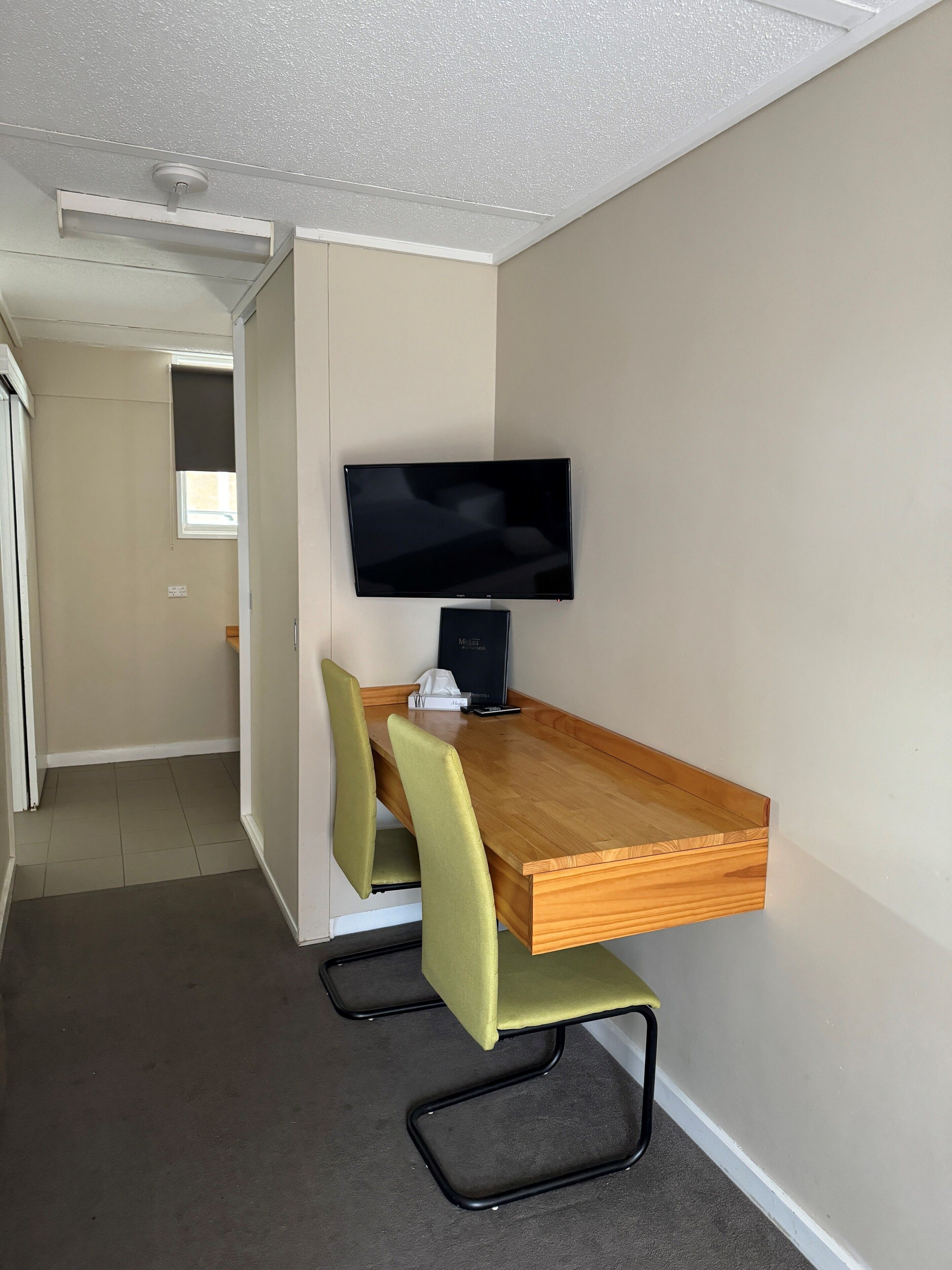 A compact room with a wooden desk, a flat-screen TV, and two green chairs.