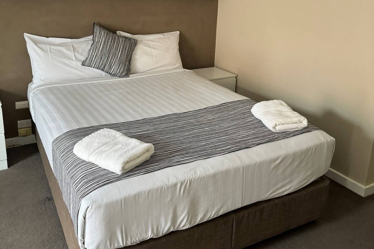 A neatly made bed with white and striped bedding, a brown headboard, and two white towels.