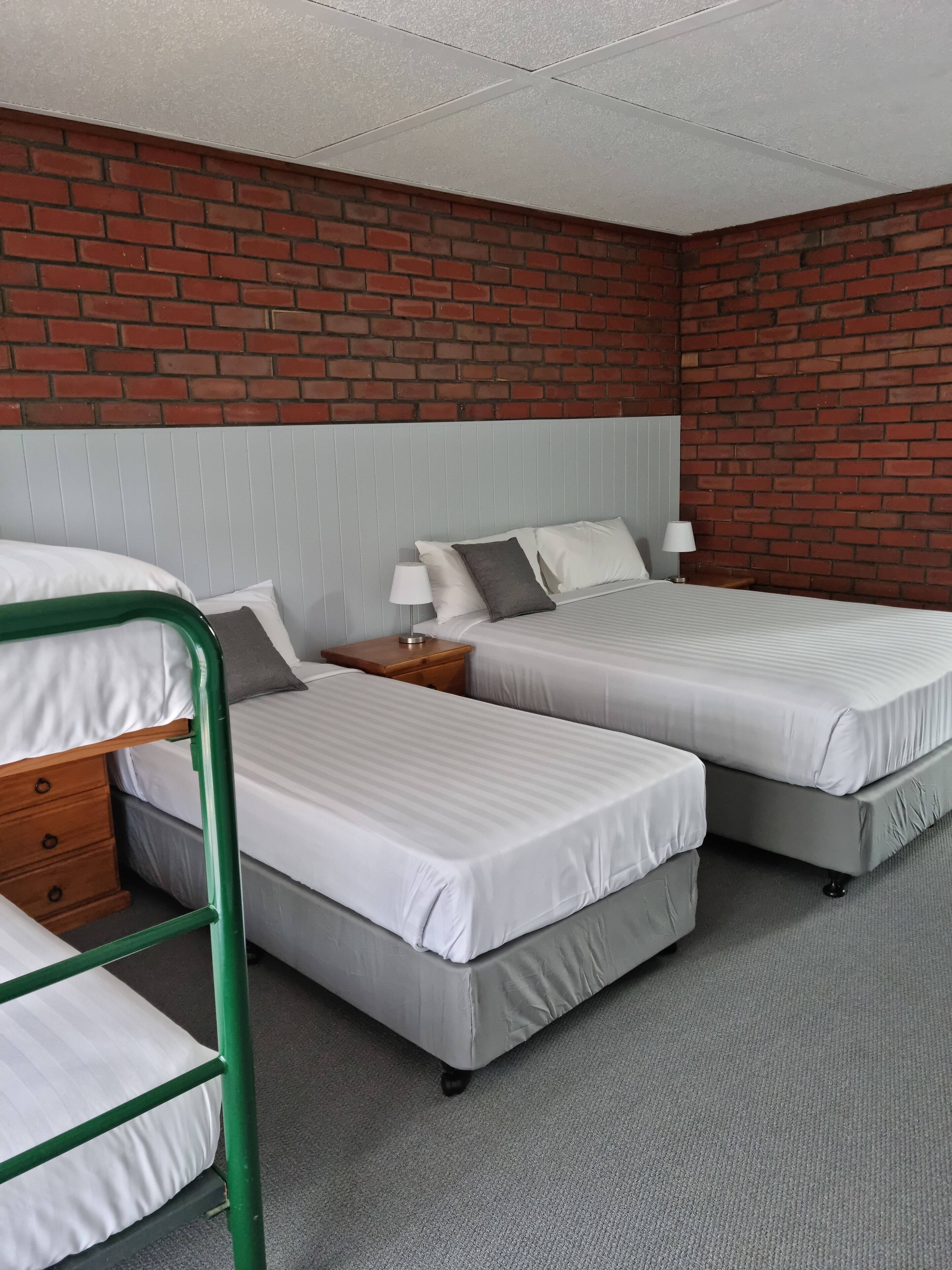 Una habitación con dos camas individuales, una cama litera de metal verde y una pared de ladrillo, además de un techo blanco.