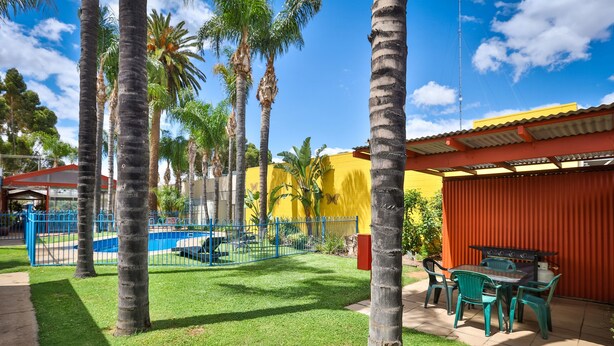 A patio area with a pool, palm trees, and a red building. - Mid City Plantation Motel (Mildura)