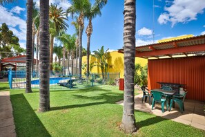 A patio area with a pool, palm trees, and a red building. - Mid City Plantation Motel (Mildura)