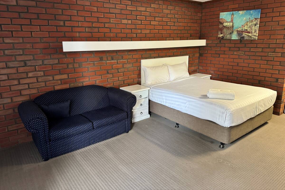 A hotel room with a brick wall, a bed with white bedding, a nightstand, a drawer unit, and a blue patterned chair.