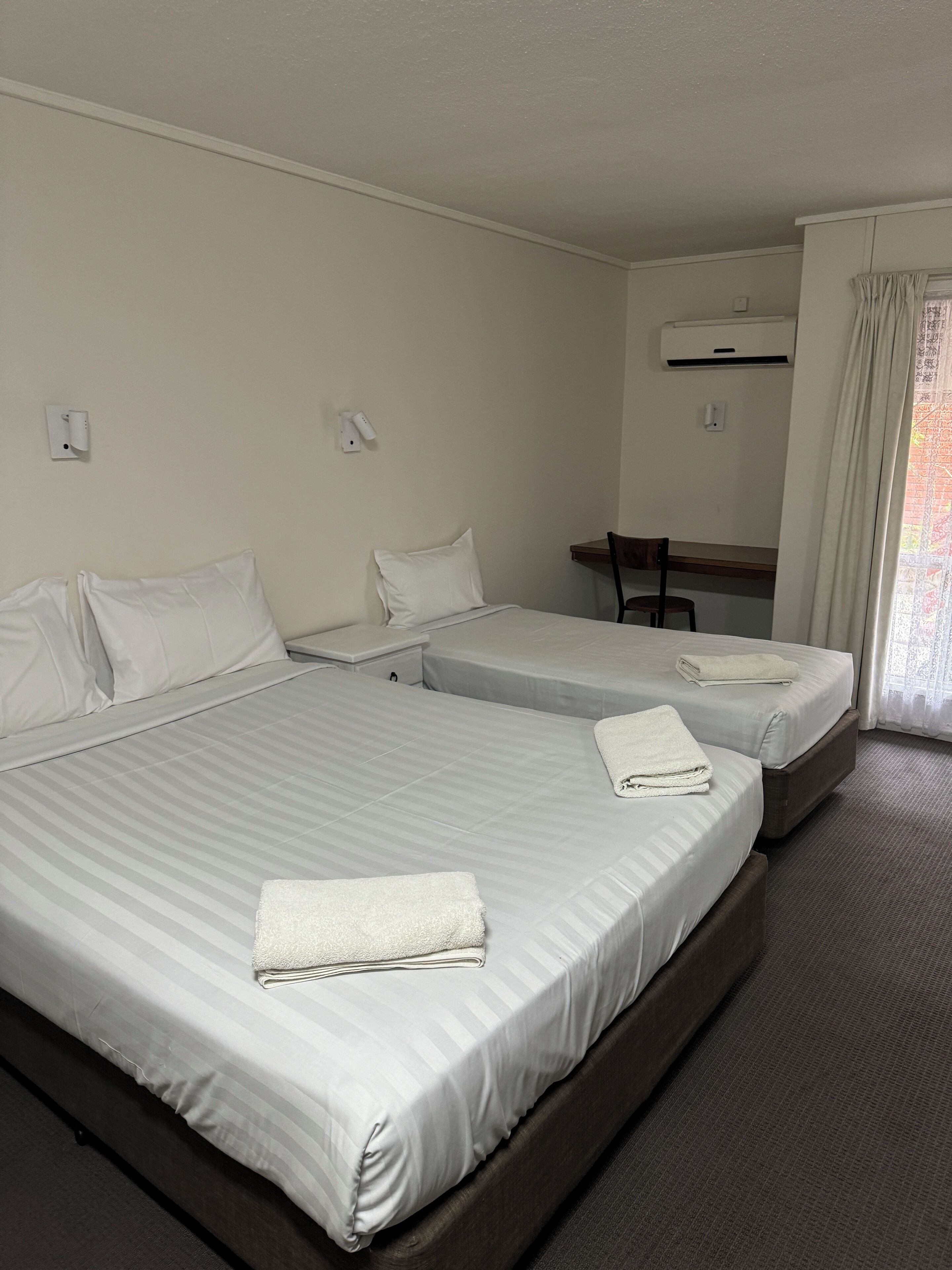 A hotel room with two beds, a desk, and an air conditioning unit.