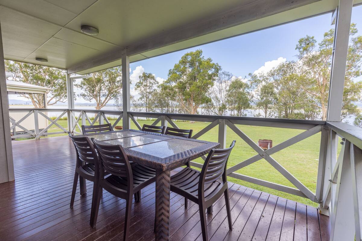 A covered outdoor area with a table and chairs, a view of a grassy field and trees, and a white railing.
