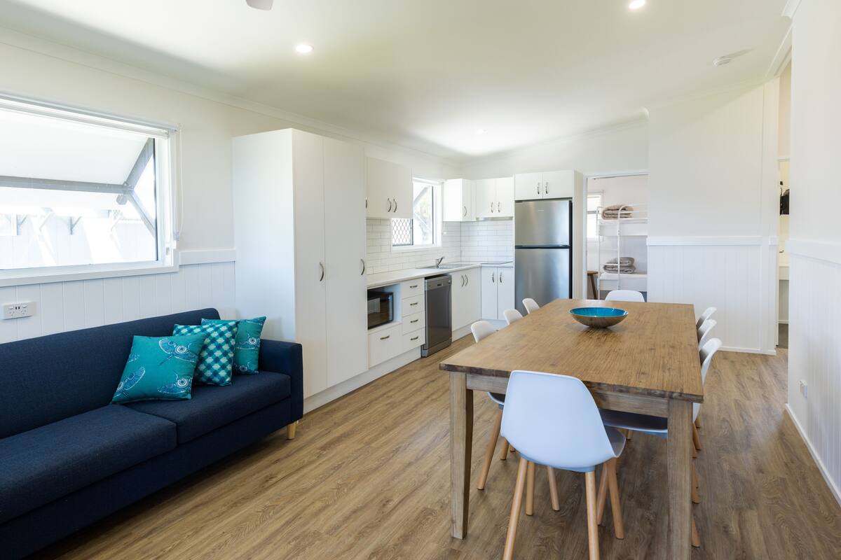A modern kitchen with white cabinets, a wooden dining table, and a blue sofa with teal cushions.