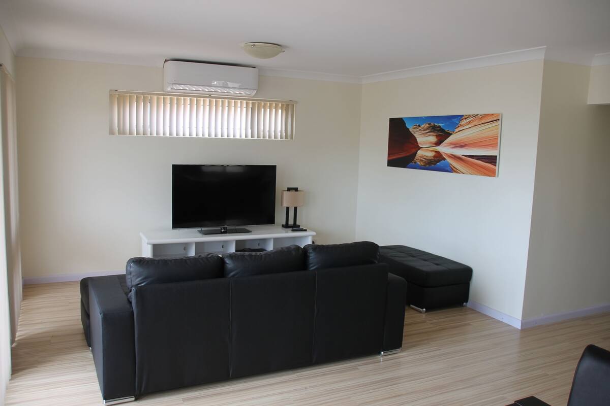 A living room with a black leather sofa, a flat-screen TV, a white entertainment unit, and a painting on the wall.
