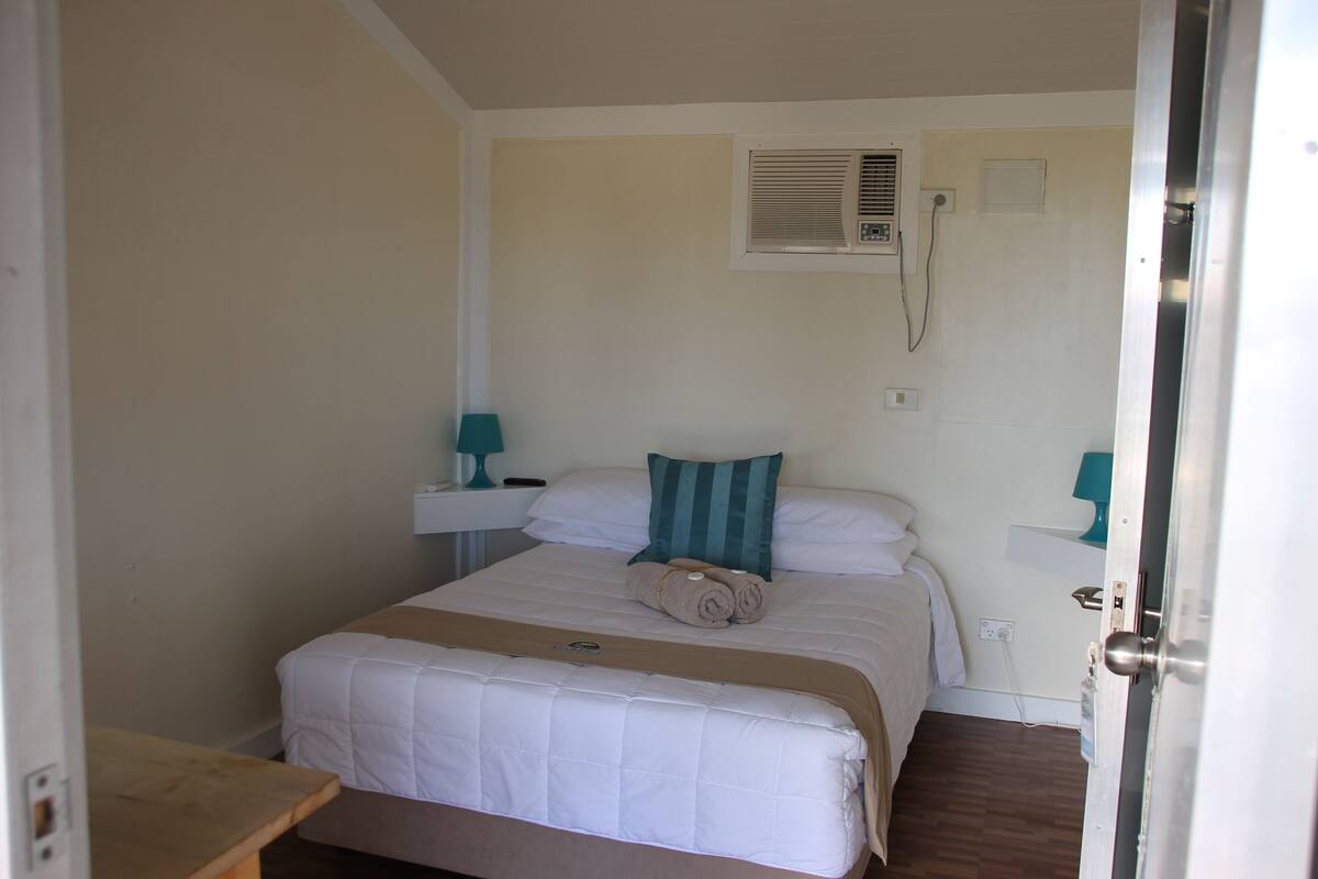 A small, neatly made bed with a white and beige duvet, a teal pillow, and a rolled towel on it. There is a small bedside table with a lamp on it.