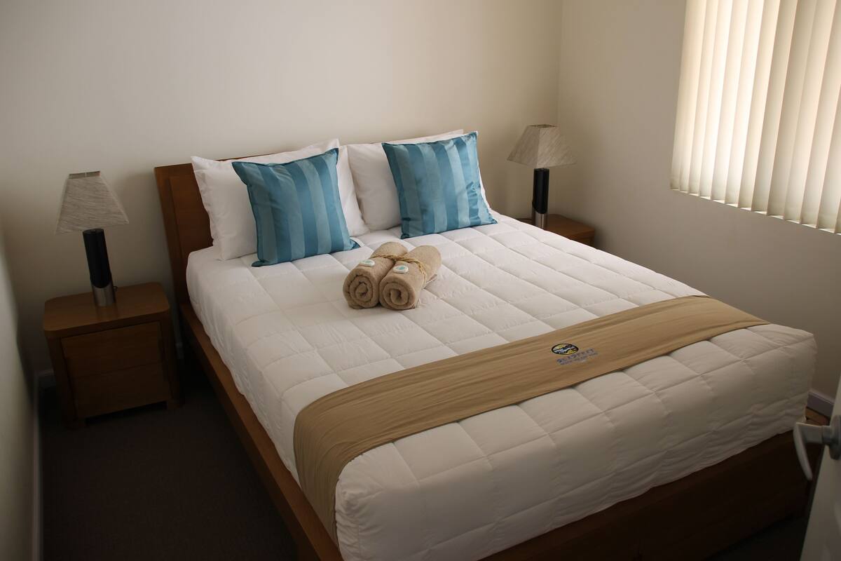 A neatly made bed with blue and white pillows, a brown blanket, and rolled towels on the side.