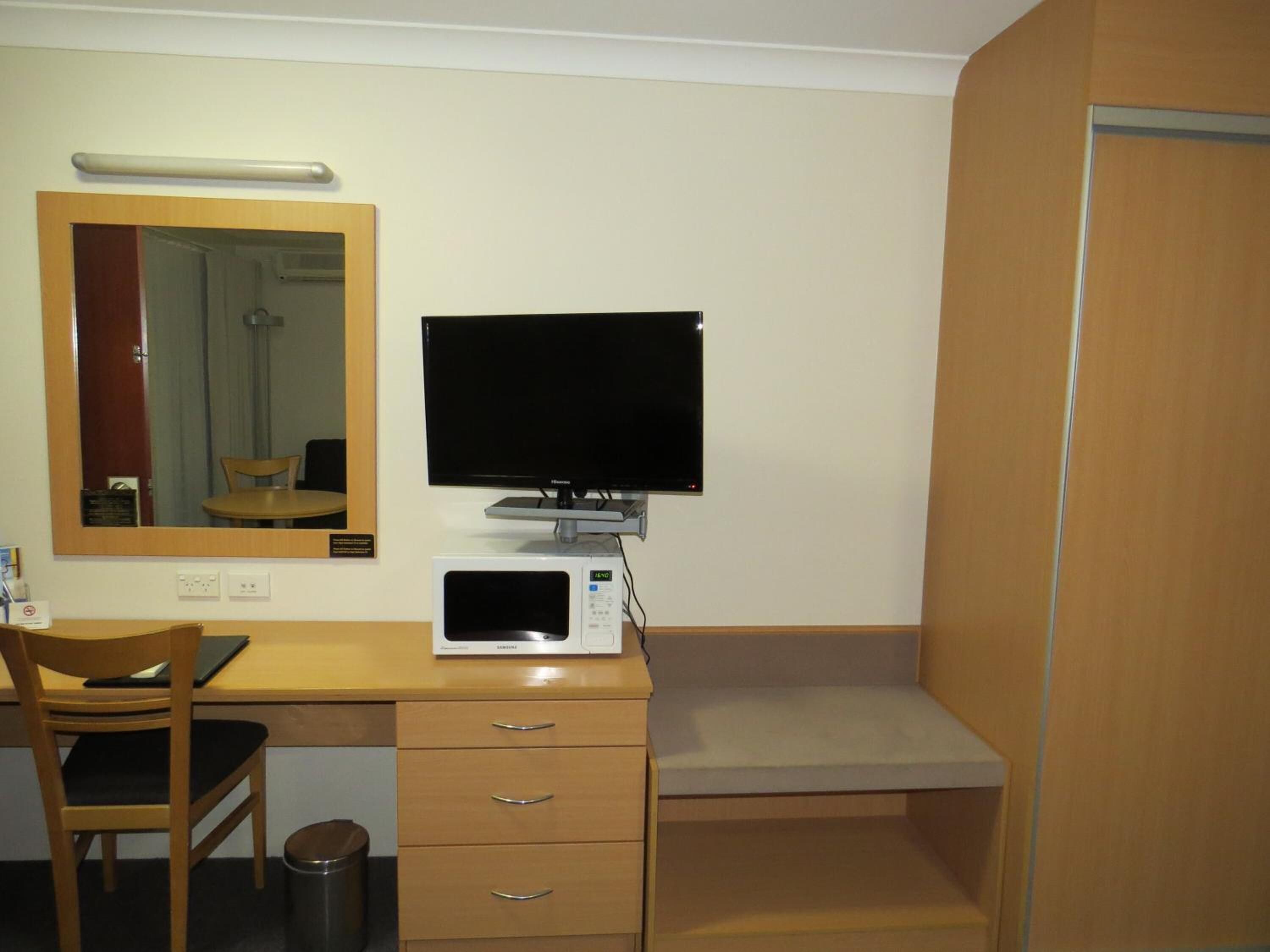 A hotel room with a wooden desk, a television, a microwave, a chair, and a trash can.