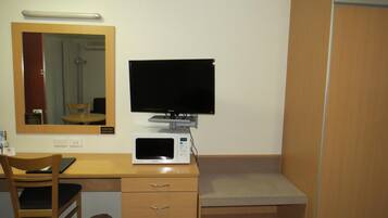 A hotel room with a wooden desk, a television, a microwave, a chair, and a trash can.