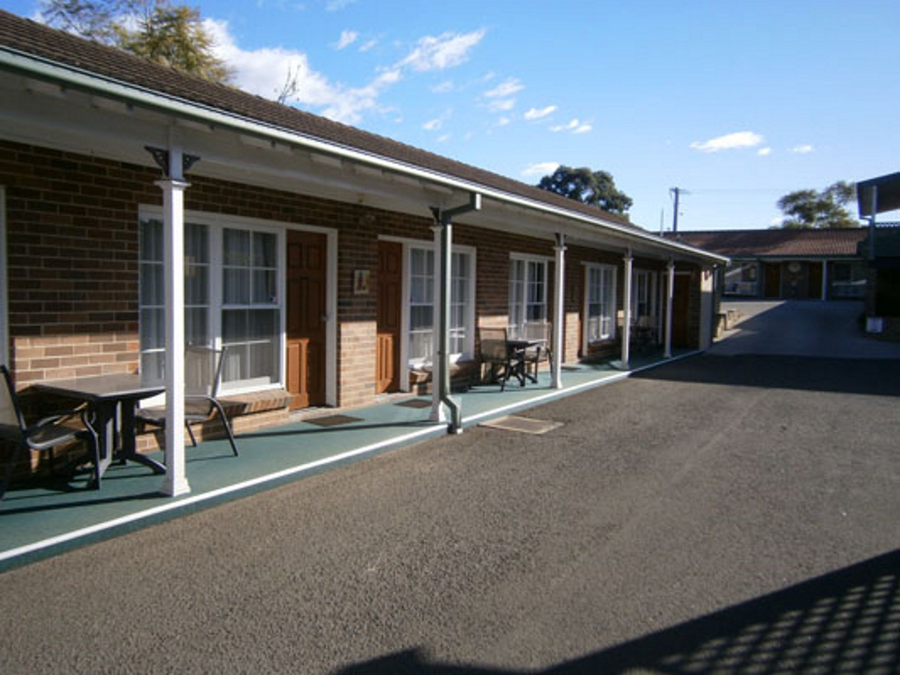 A motel with a covered outdoor seating area, individual rooms with windows, and a paved parking area.