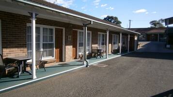 A motel with a covered outdoor seating area, individual rooms with windows, and a paved parking area.