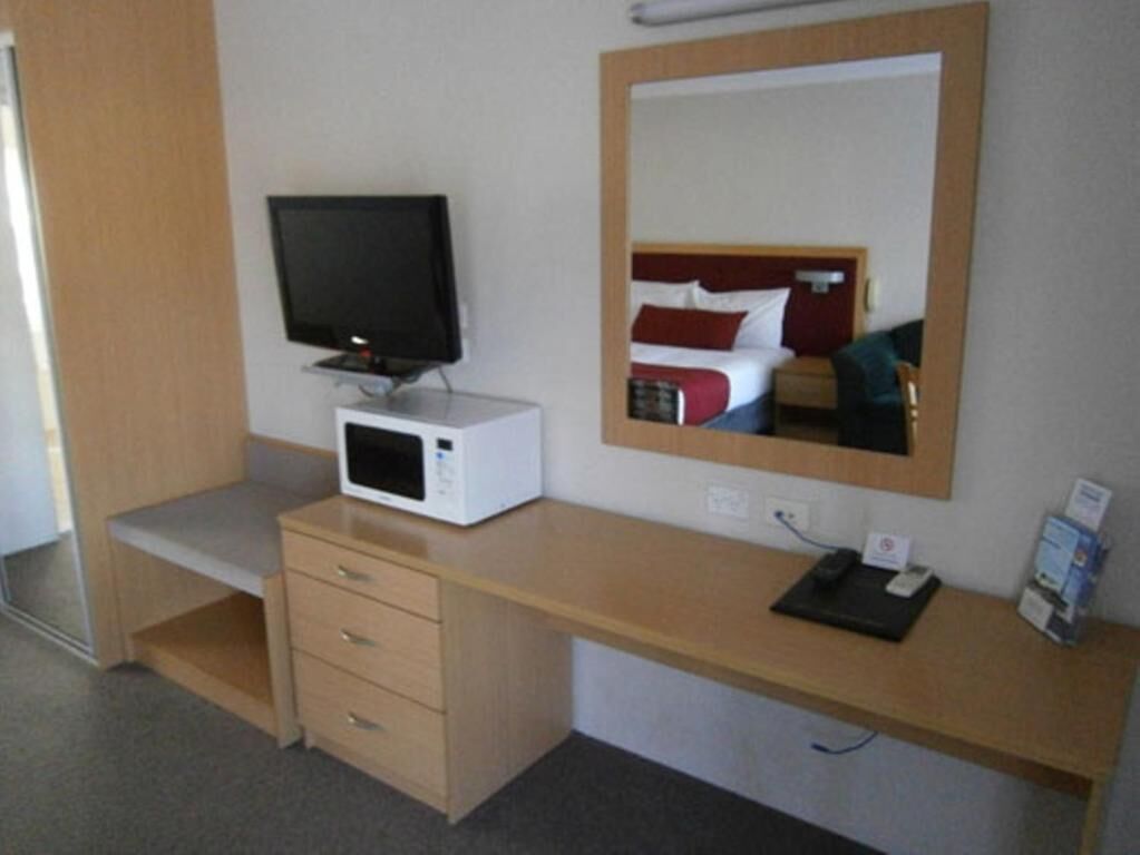 Habitación de hotel con escritorio de madera, televisión de pantalla plana, microondas, espejo y cama con ropa de cama roja y blanca.