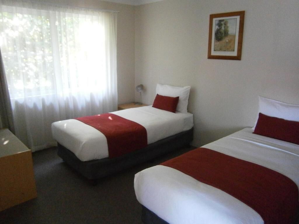 Habitación de hotel con dos camas, una mesita de noche, una lámpara y una ventana con cortinas traslúcidas.