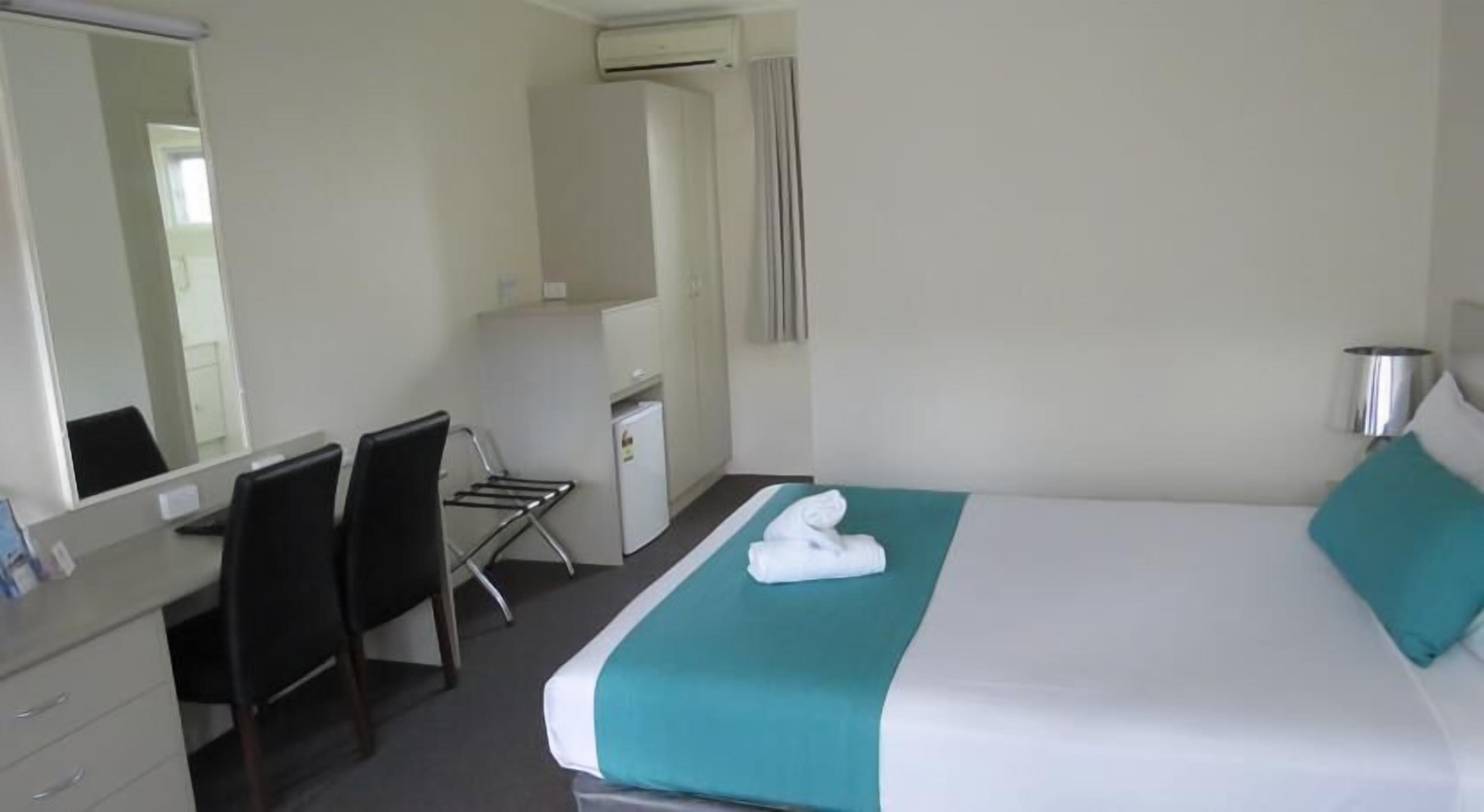 Habitación de hotel con cama, escritorio, silla, espejo y un refrigerador pequeño.