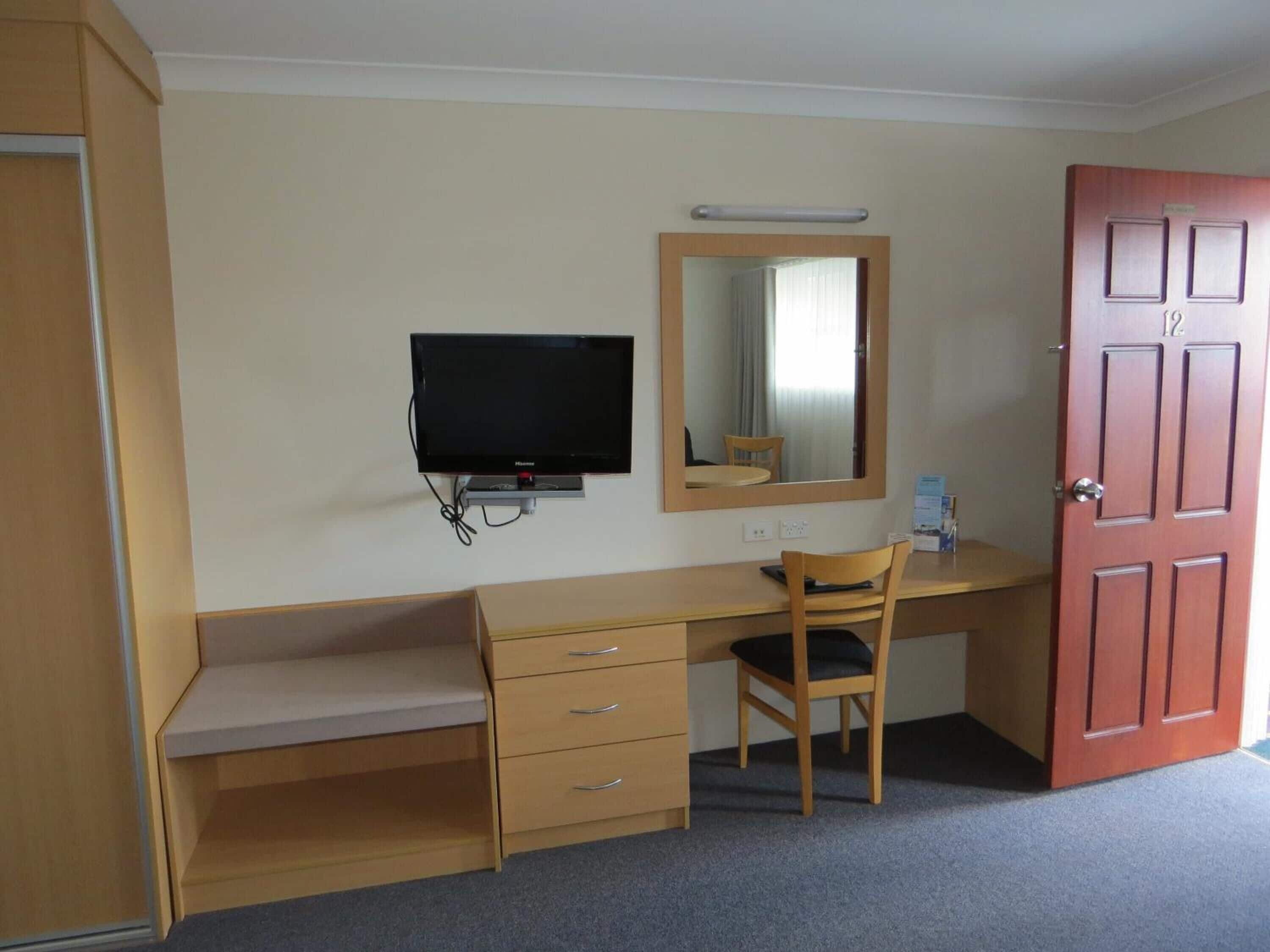 A hotel room with a wooden desk, a television, a mirror, and a door.