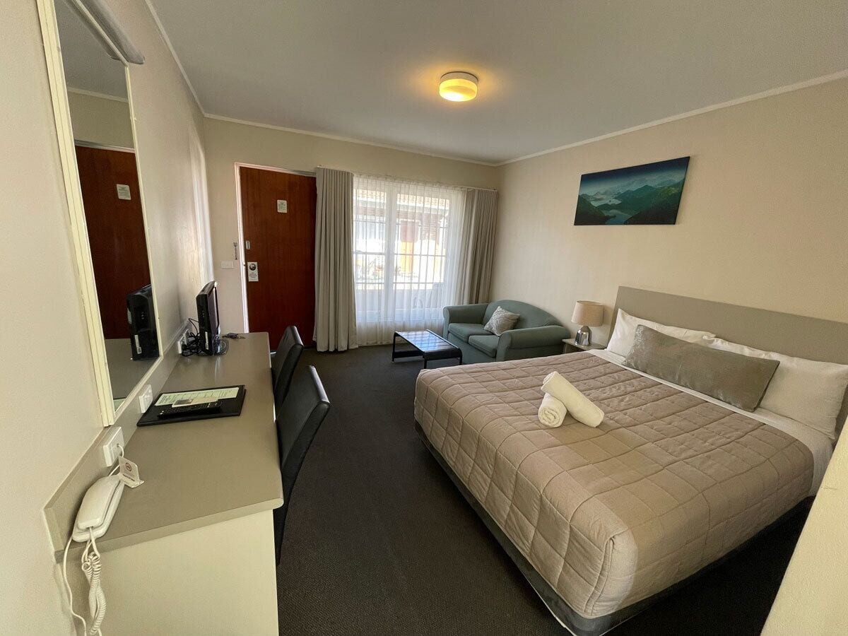 Habitación de hotel con una cama, un escritorio con teléfono, una silla, una mesita y una ventana con cortinas.