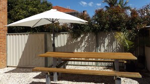 Una mesa de picnic de madera con un paraguas blanco en un patio trasero con cerca blanca, suelo de grava y árboles.