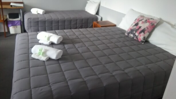Habitación de hotel con dos camas, cada una con cubrecama gris acolchado y almohadas blancas. Hay dos toallas enrolladas sobre la cama. Una mesita de noche de madera con una lámpara está entre las camas.