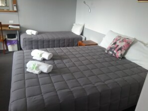 A hotel room with two beds, each with a grey quilted bedspread and white pillows. There are two rolled towels on the bed. A small wooden nightstand with a lamp is between the beds. - Civic Motel (Grafton)