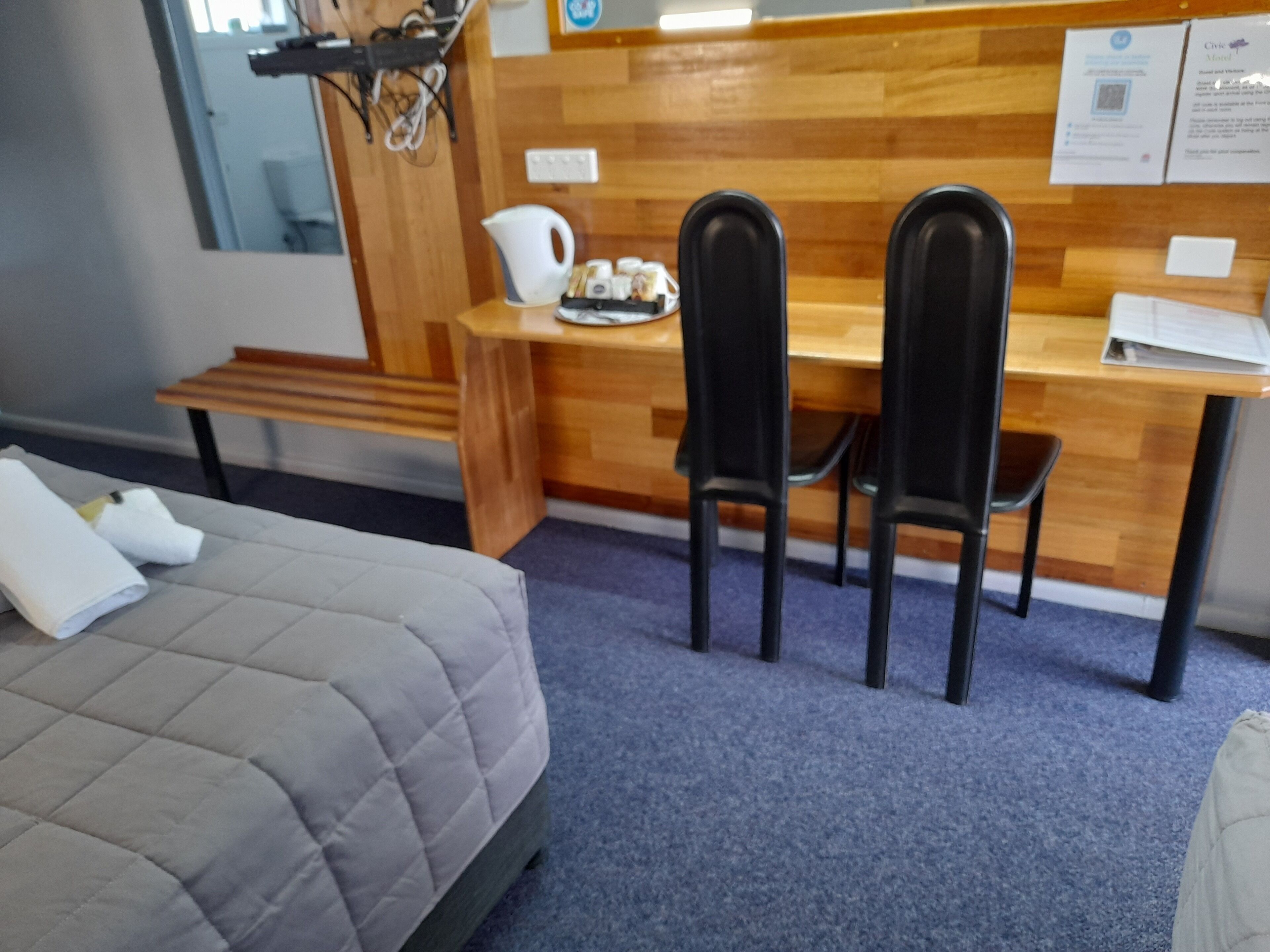 Une chambre d’hôtel comprenant un lit, un bureau, deux chaises, une petite table avec une carafe et des en-cas, ainsi qu’un tableau d’affichage avec des documents.