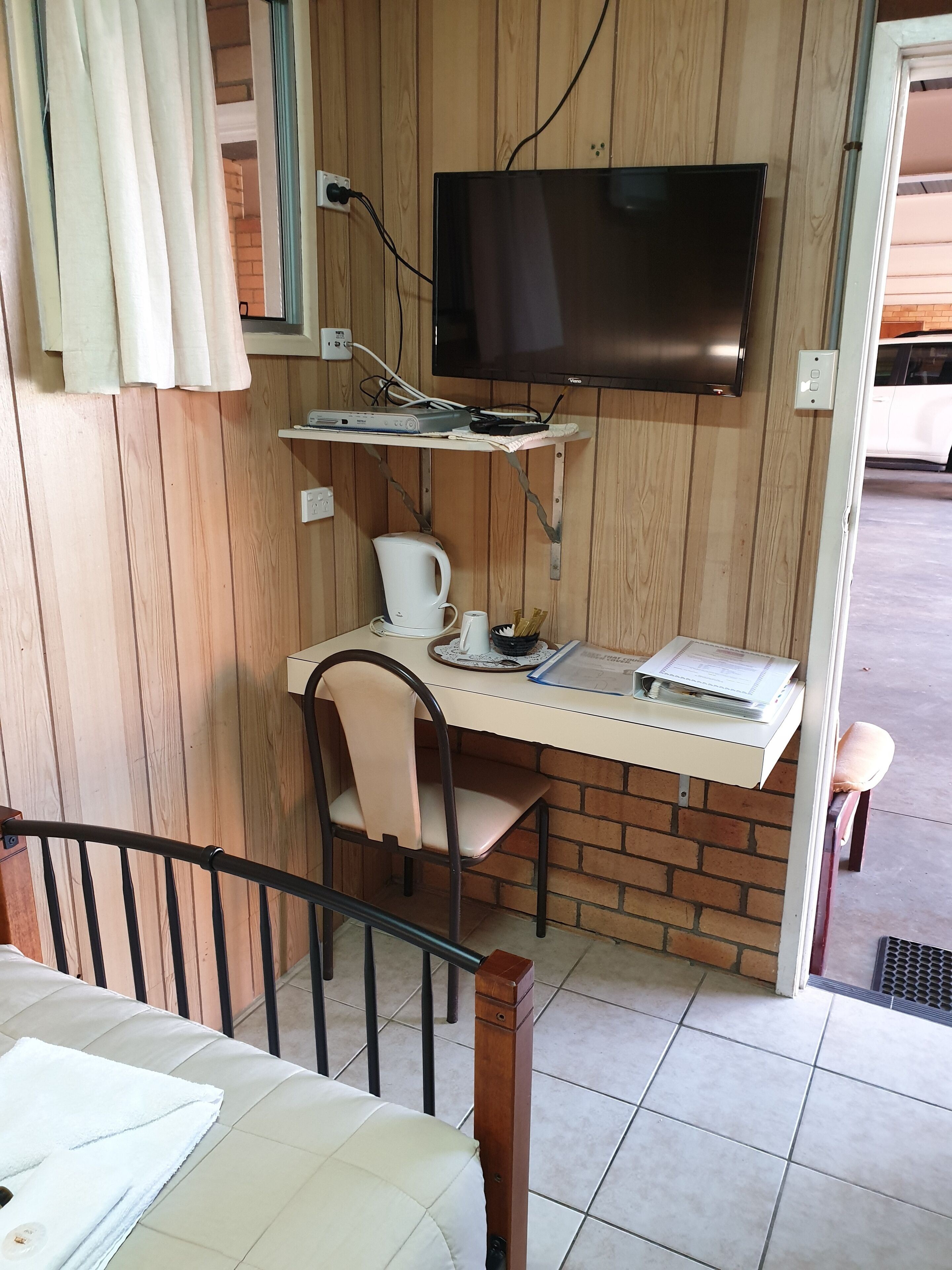 Quarto compacto com televisão na parede, uma secretária pequena com cadeira, chaleira e uma chávena.