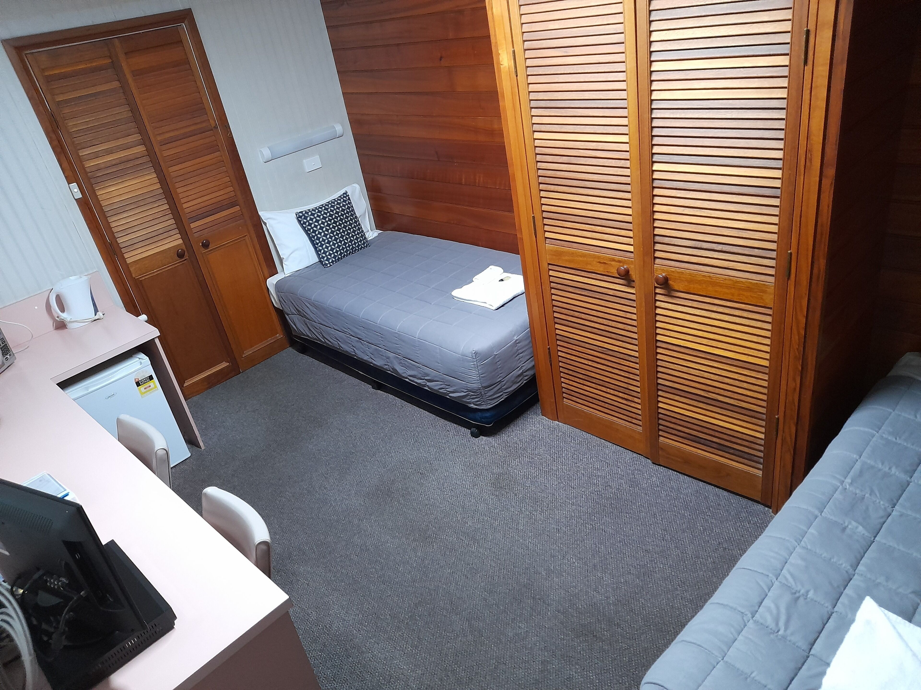 A compact room with a bed, a desk with a computer, a small refrigerator, and a wardrobe.