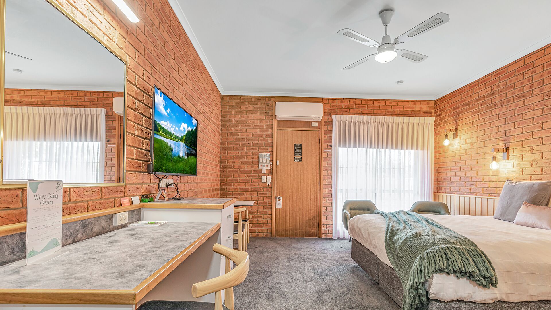 Une chambre d’hôtel avec un mur en briques, un lit, un bureau avec une chaise, une télévision et un ventilateur de plafond.