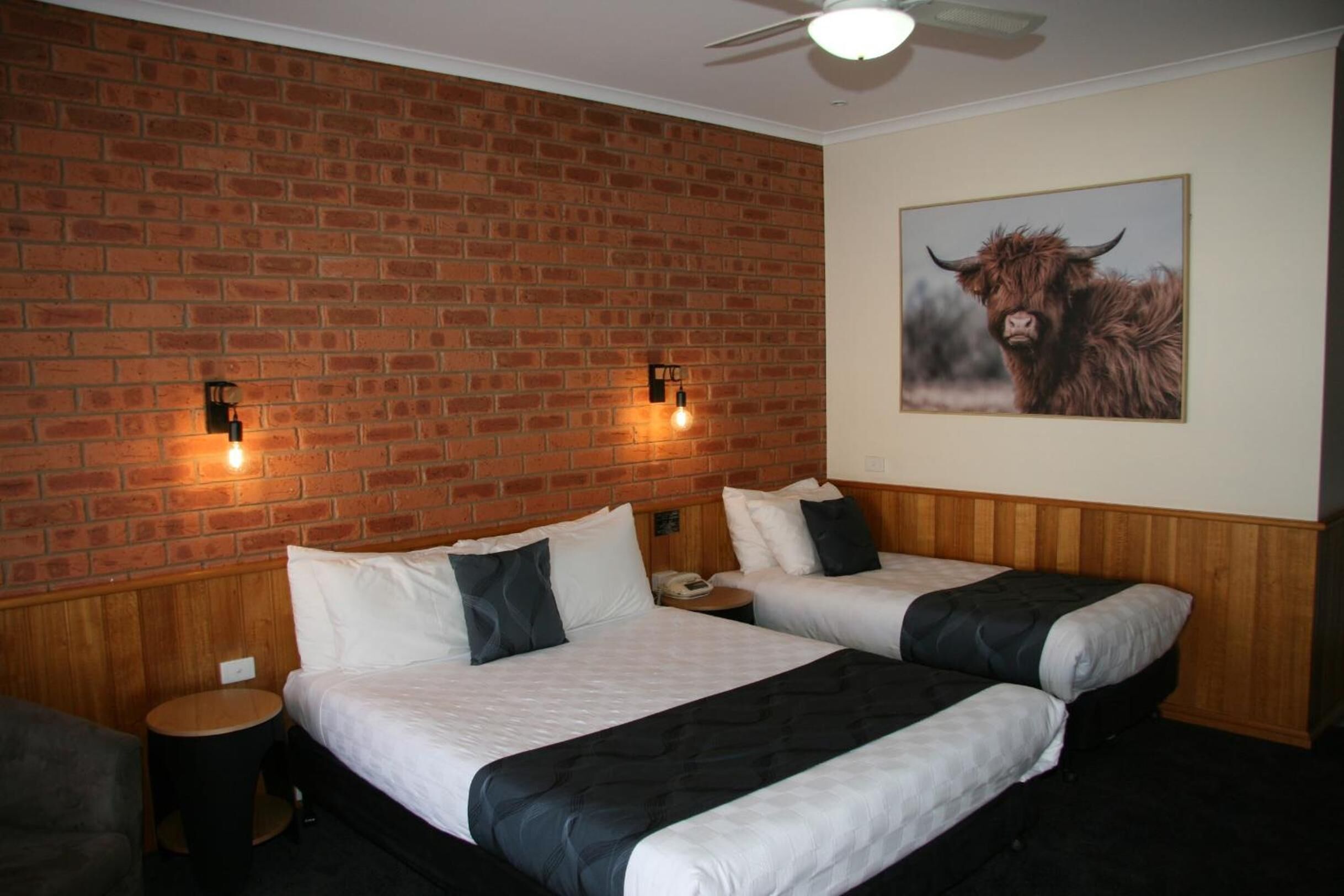 A hotel room with two beds, a brick wall, a ceiling fan, and a painting of a bull.