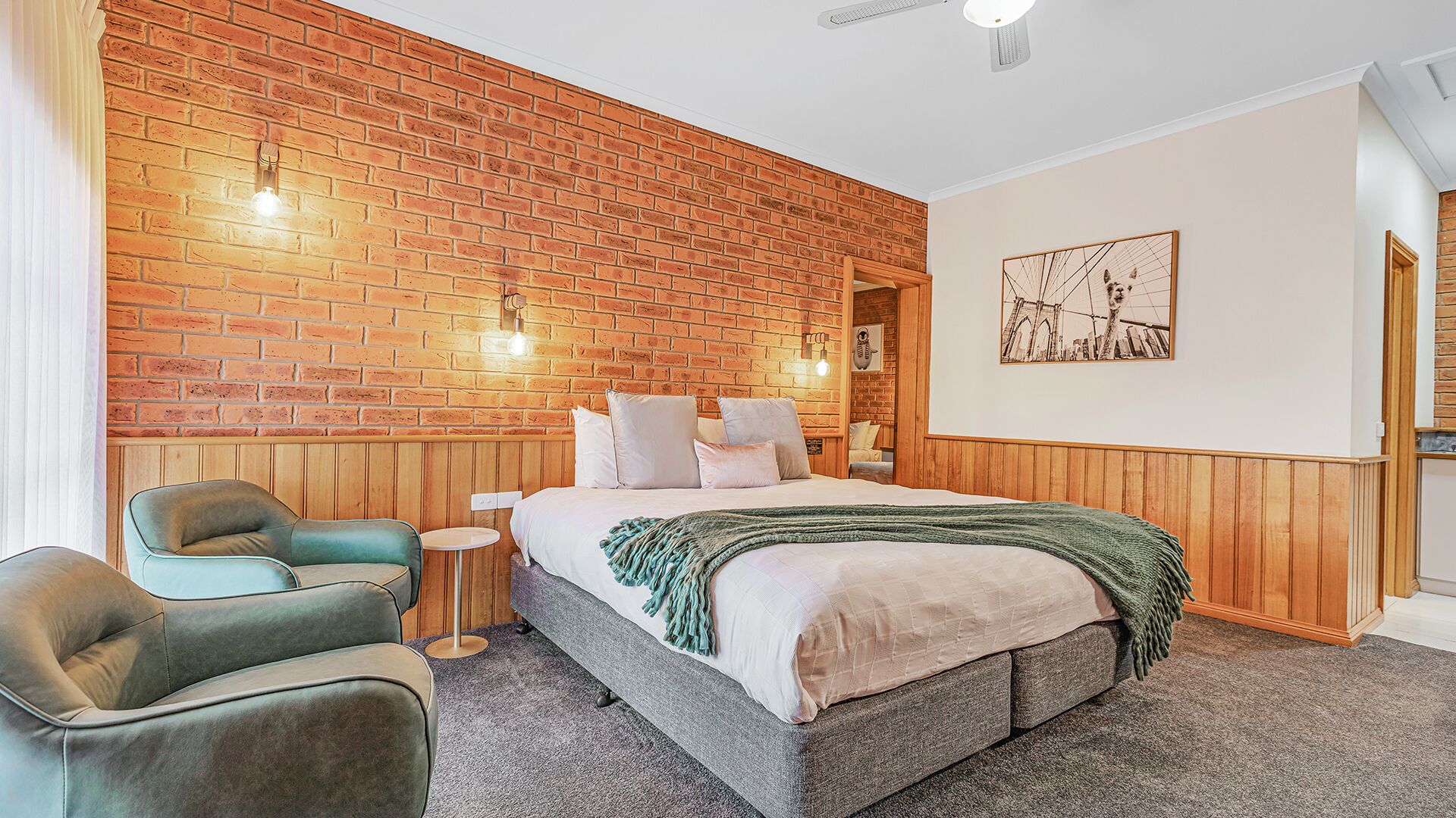 A bedroom with a brick wall, a bed with pillows, a green armchair, a small round table, and a painting on the wall.