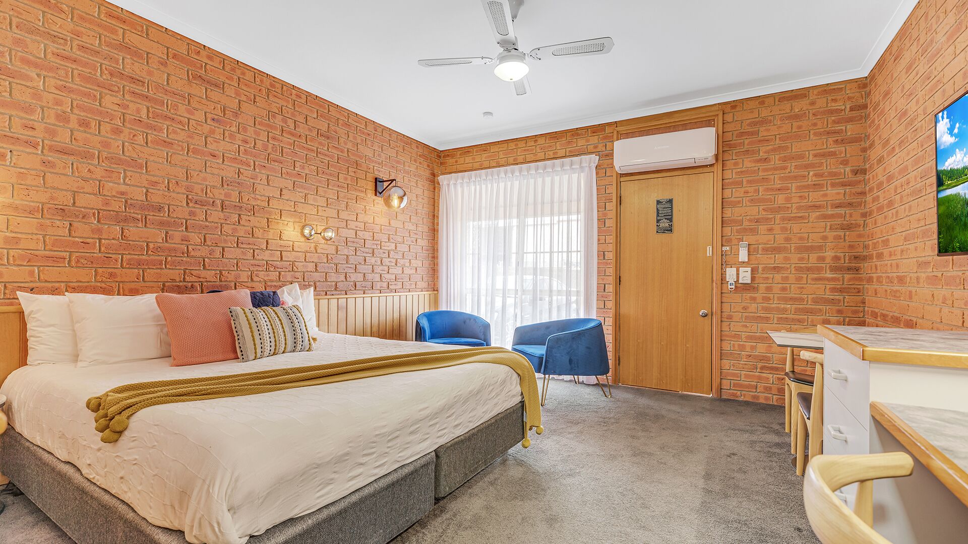 Un dormitorio con pared de ladrillo, una cama con edredón blanco, ventilador de techo y una ventana con cortinas.