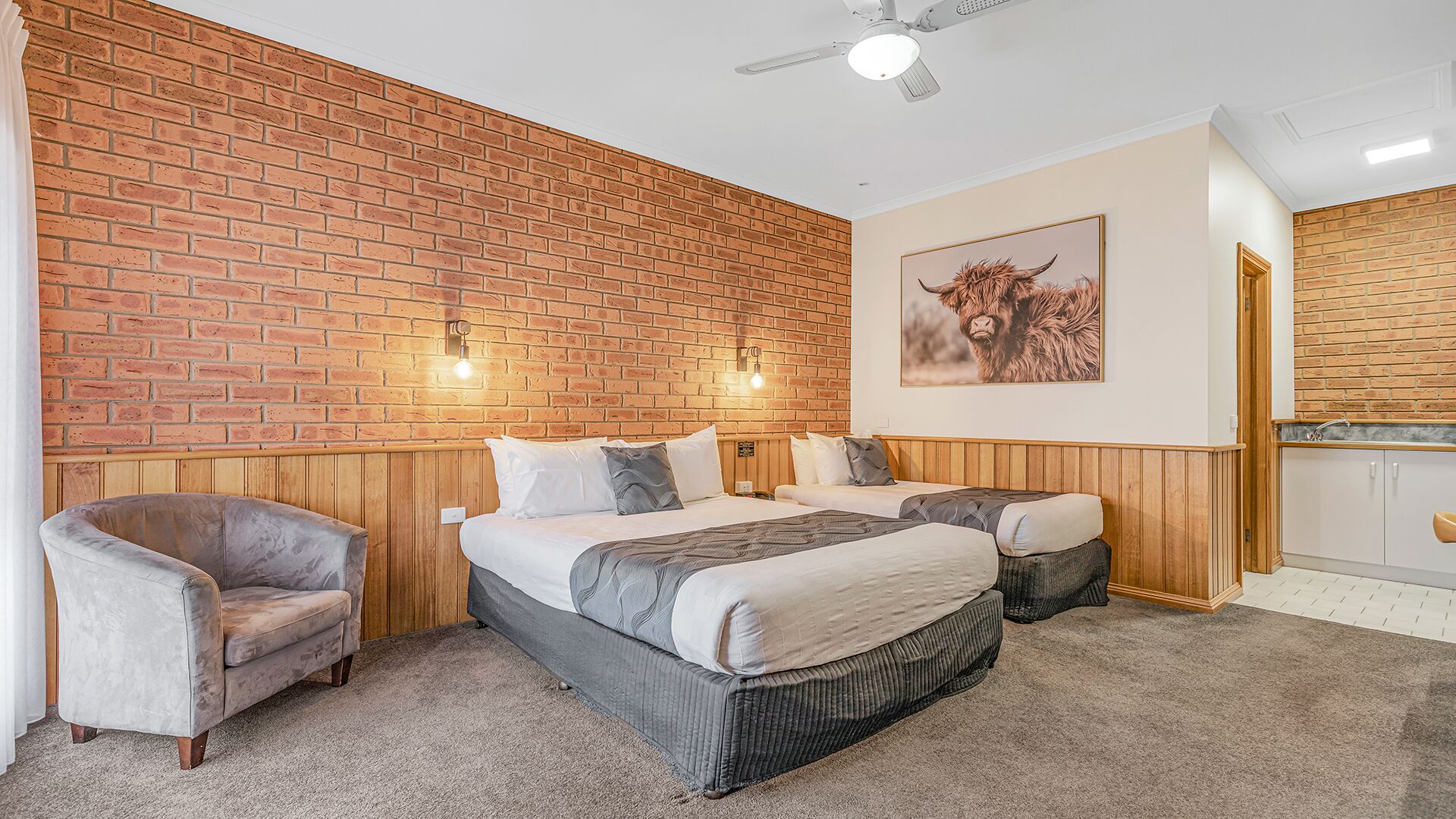 Una habitación con pared de ladrillo, una cama con dos almohadas, una silla y un cuadro en la pared.