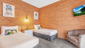 A room with two beds, a wooden nightstand, a leather armchair, and a brick wall with artwork.