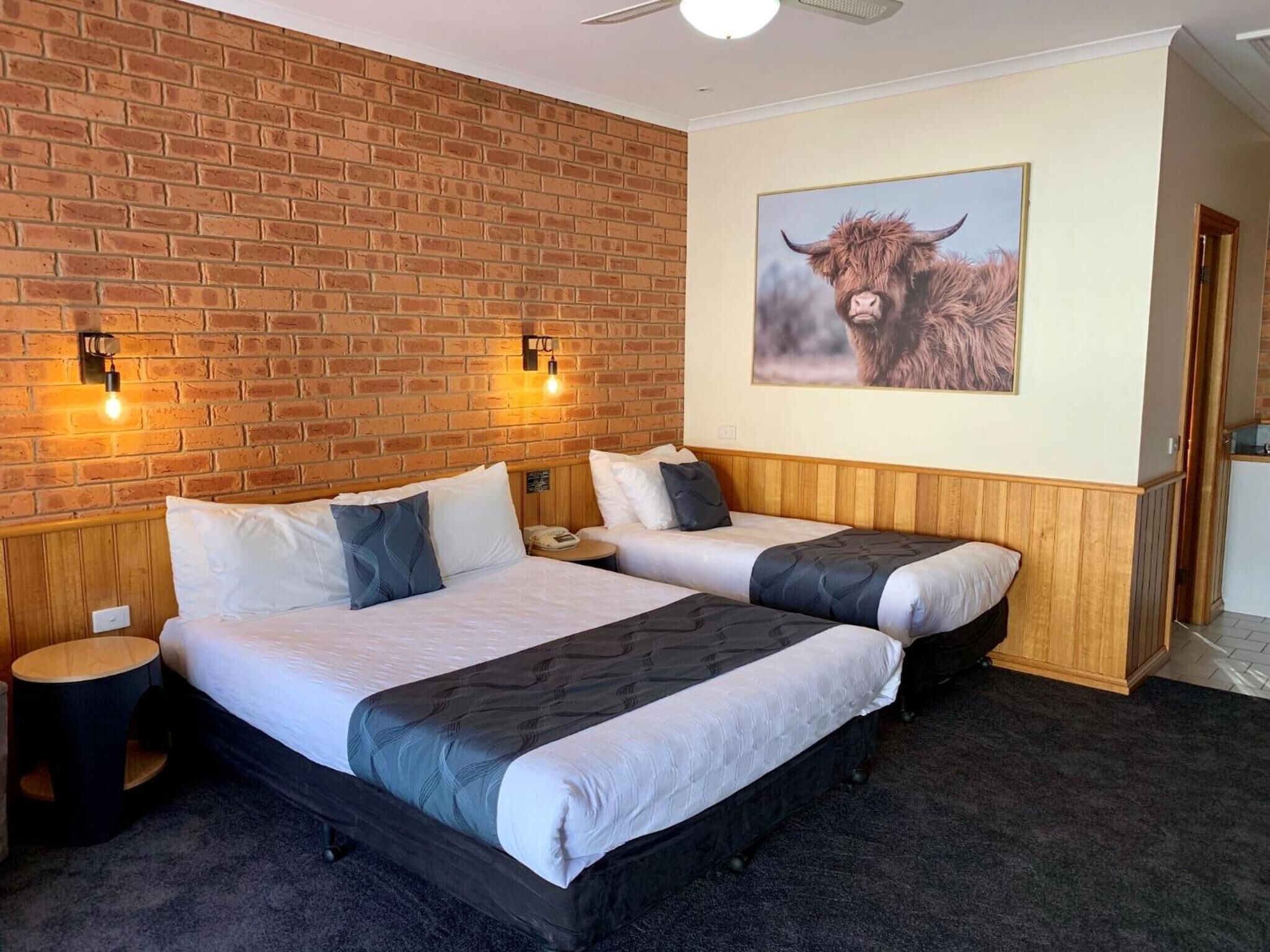 Habitación de hotel con dos camas, pared de ladrillo, cuadro colgado de un toro y cabecero de madera.