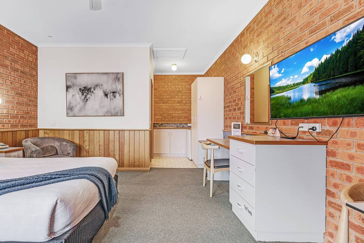 A hotel room with a bed, a desk, a television, a painting, and a brick wall.