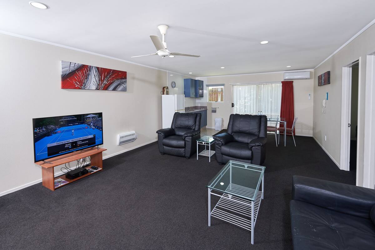 A modern living room with a flat-screen TV, leather sofas, a glass coffee table, and a ceiling fan.
