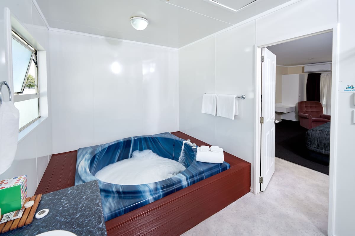 A modern bathroom with a large bathtub, a shower area, and a toilet. There is a towel rack with two towels, and a small table with a bottle and a tissue box.