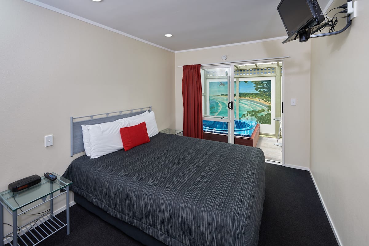 A hotel room with a bed, a glass table with a remote control, a TV mounted on the wall, and a view of a pool and landscape through a glass door.
